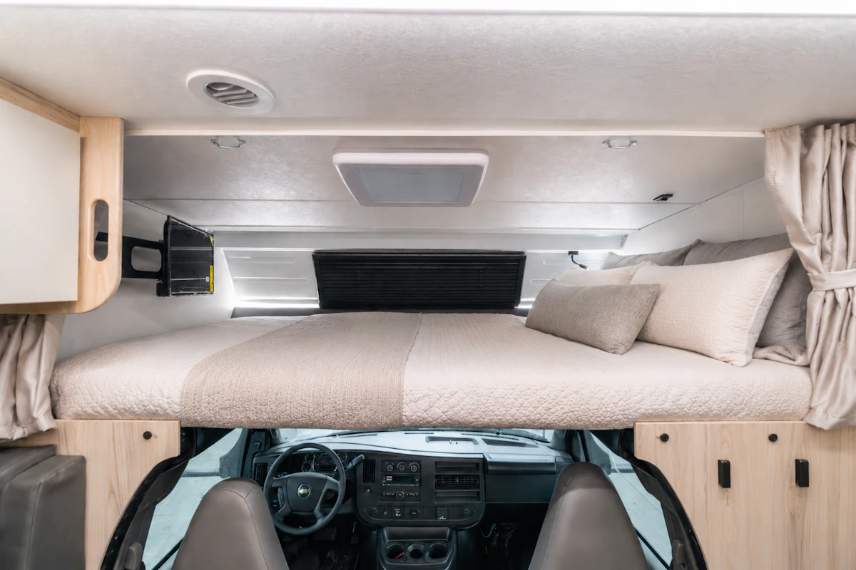 Over-cab bed area above front seats with bedding, pillows, side cabinets, small TV, and curtain dividers.