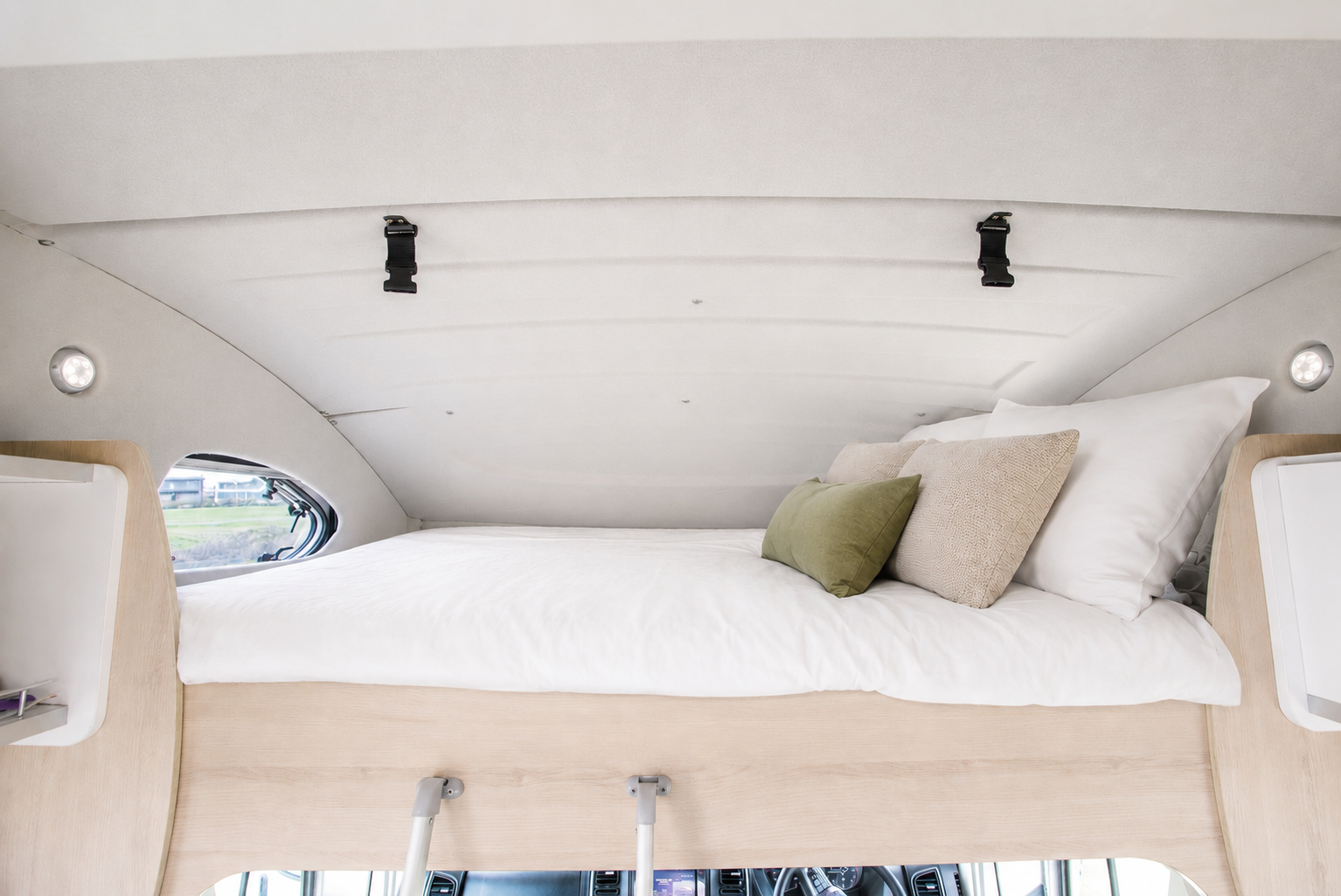 Upper bed inside a campervan with pillows, wooden frame, side shelves, and small round windows.