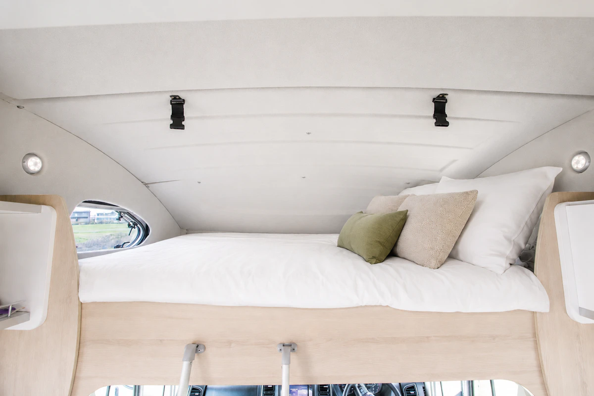 Upper bed inside a campervan with pillows, wooden frame, side shelves, and small round windows.