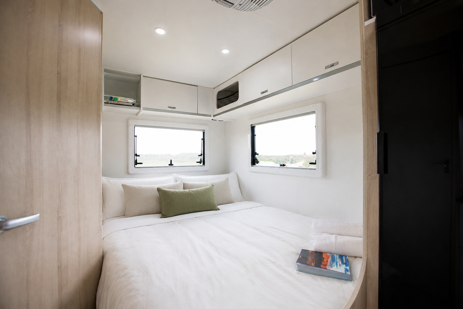 Van interior with bed, overhead cabinets, two windows, reading light, and book on bed.