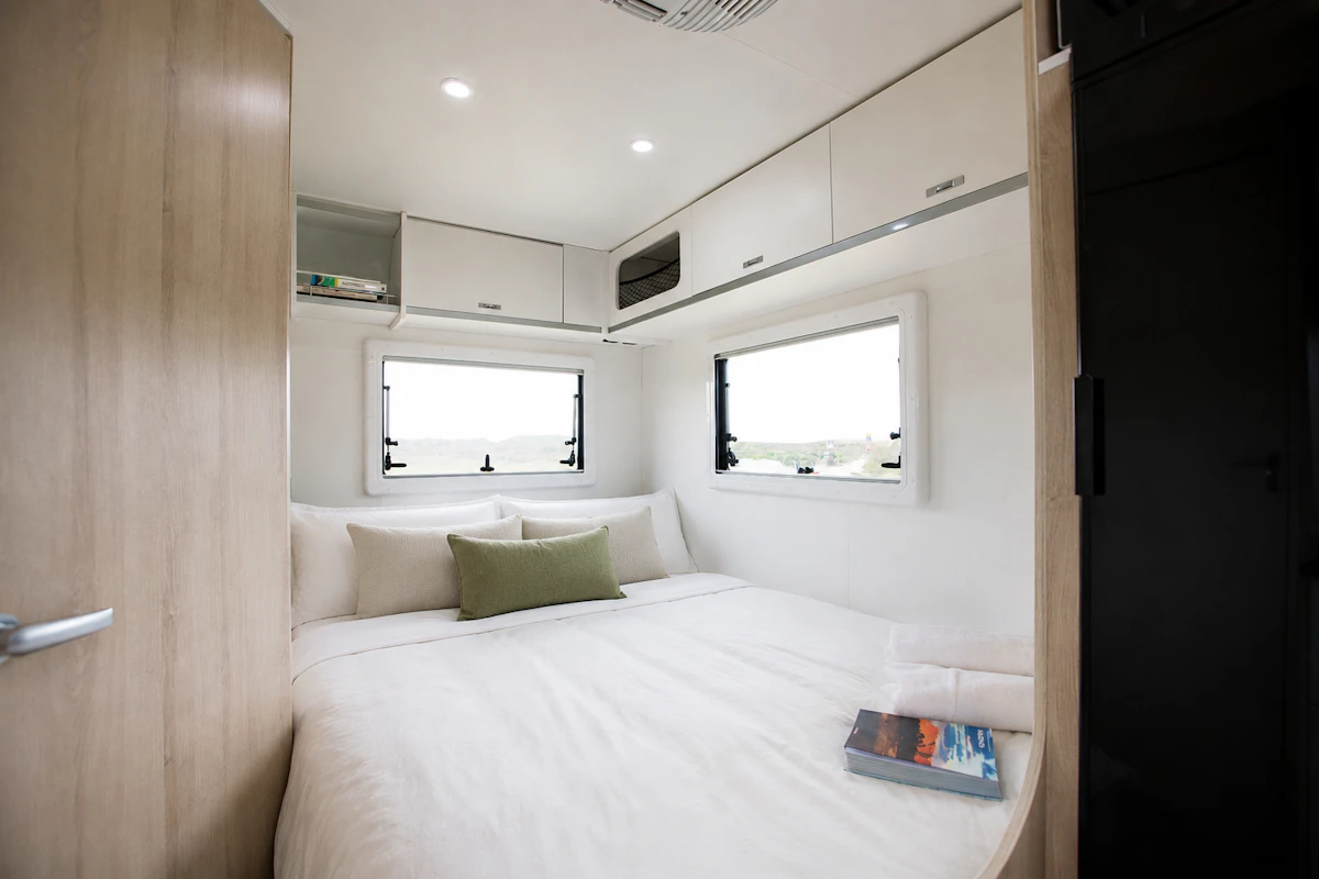 Van interior with bed, overhead cabinets, two windows, reading light, and book on bed.