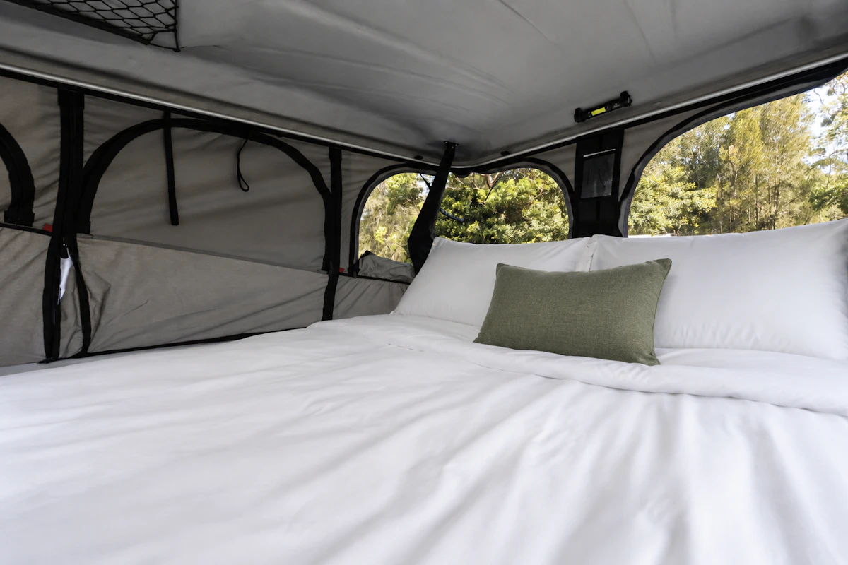 Pop-up roof tent interior with made bed, two pillows, mesh storage, zippered windows, and fabric wall panels.