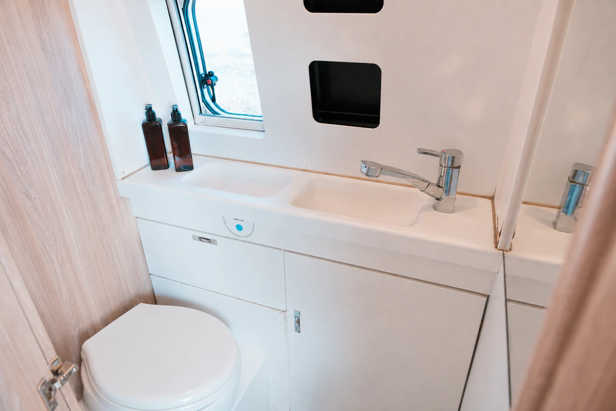 Van bathroom with toilet, sink, faucet, two bottles, window, and built-in shelf, viewed from the door.