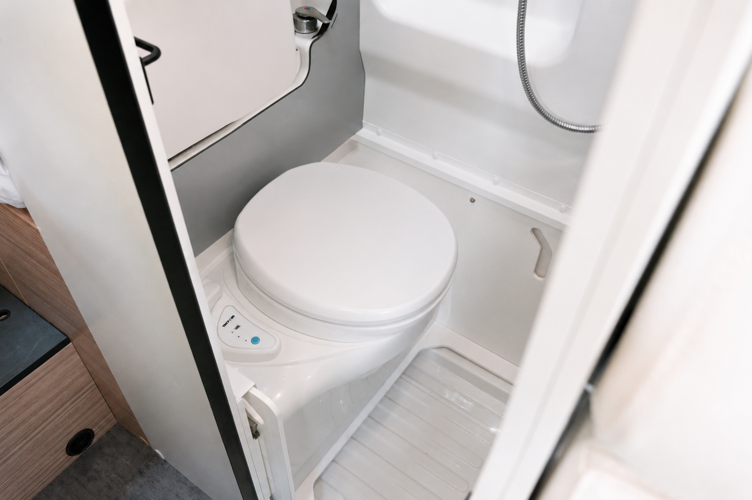 Compact camper van bathroom with a toilet, showerhead, plastic flooring, and light-colored walls visible.