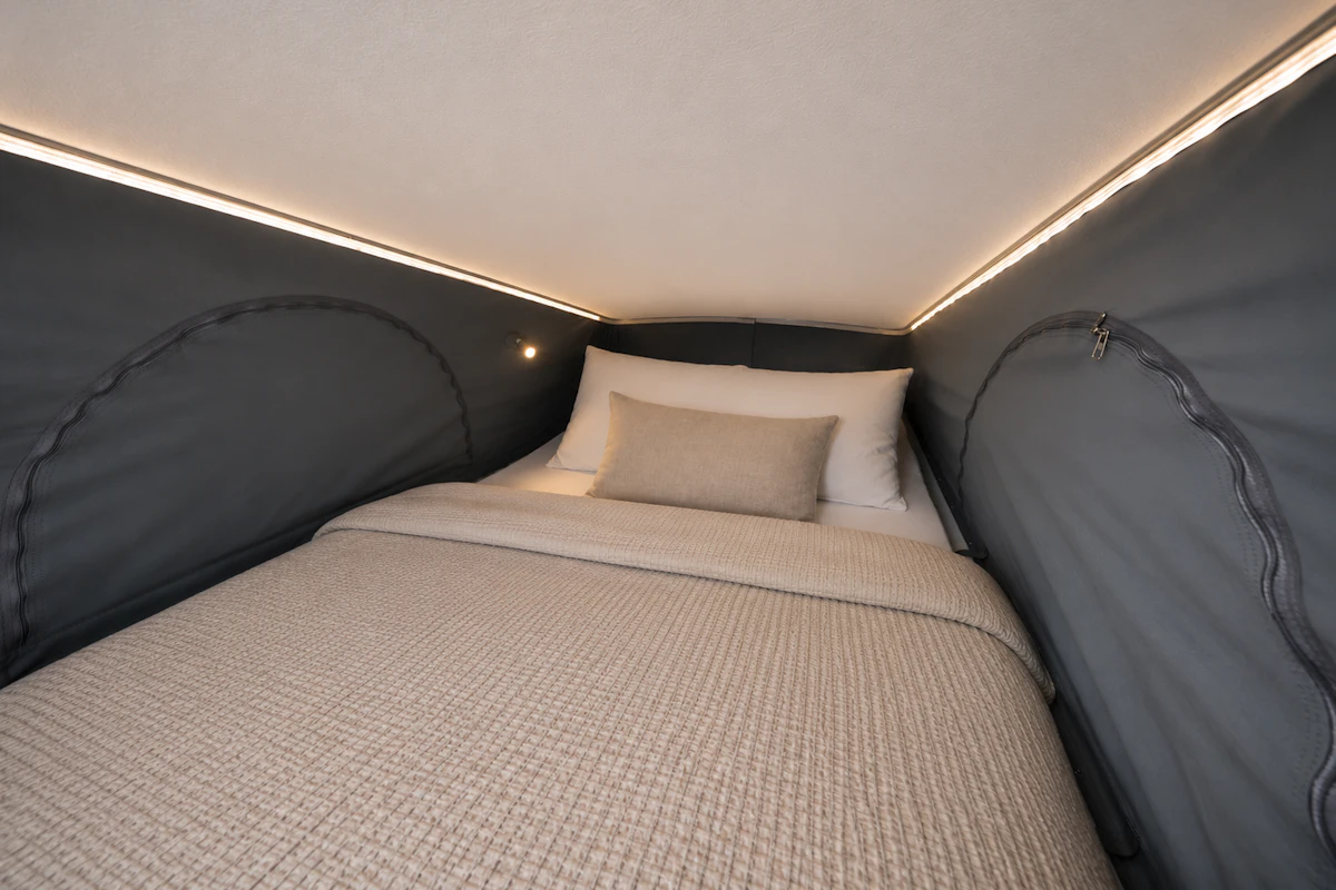 Interior van view showing bed with beige blanket and pillows under a pop-up roof tent with LED lighting.