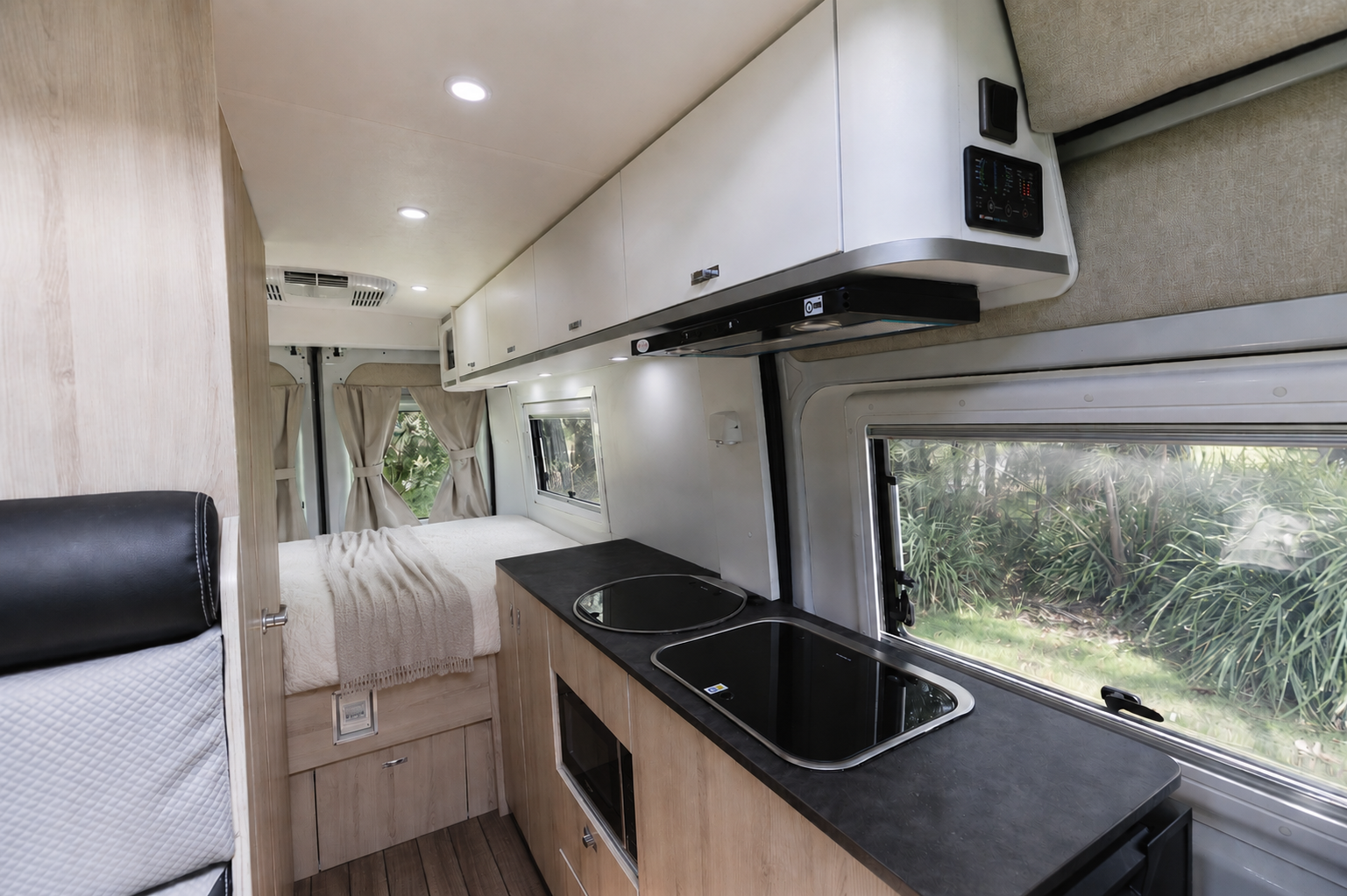 Van interior with bed, kitchenette with sink and stove, overhead cabinets, and side window.