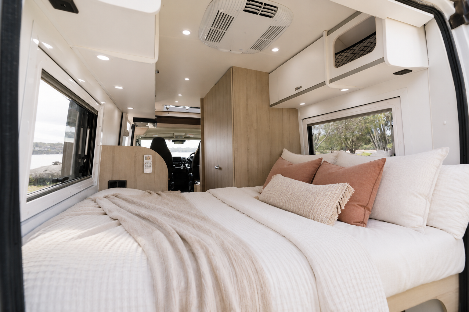 Van interior with large rear bed, overhead storage cabinets, side windows, wood paneling, and ceiling lights visible.