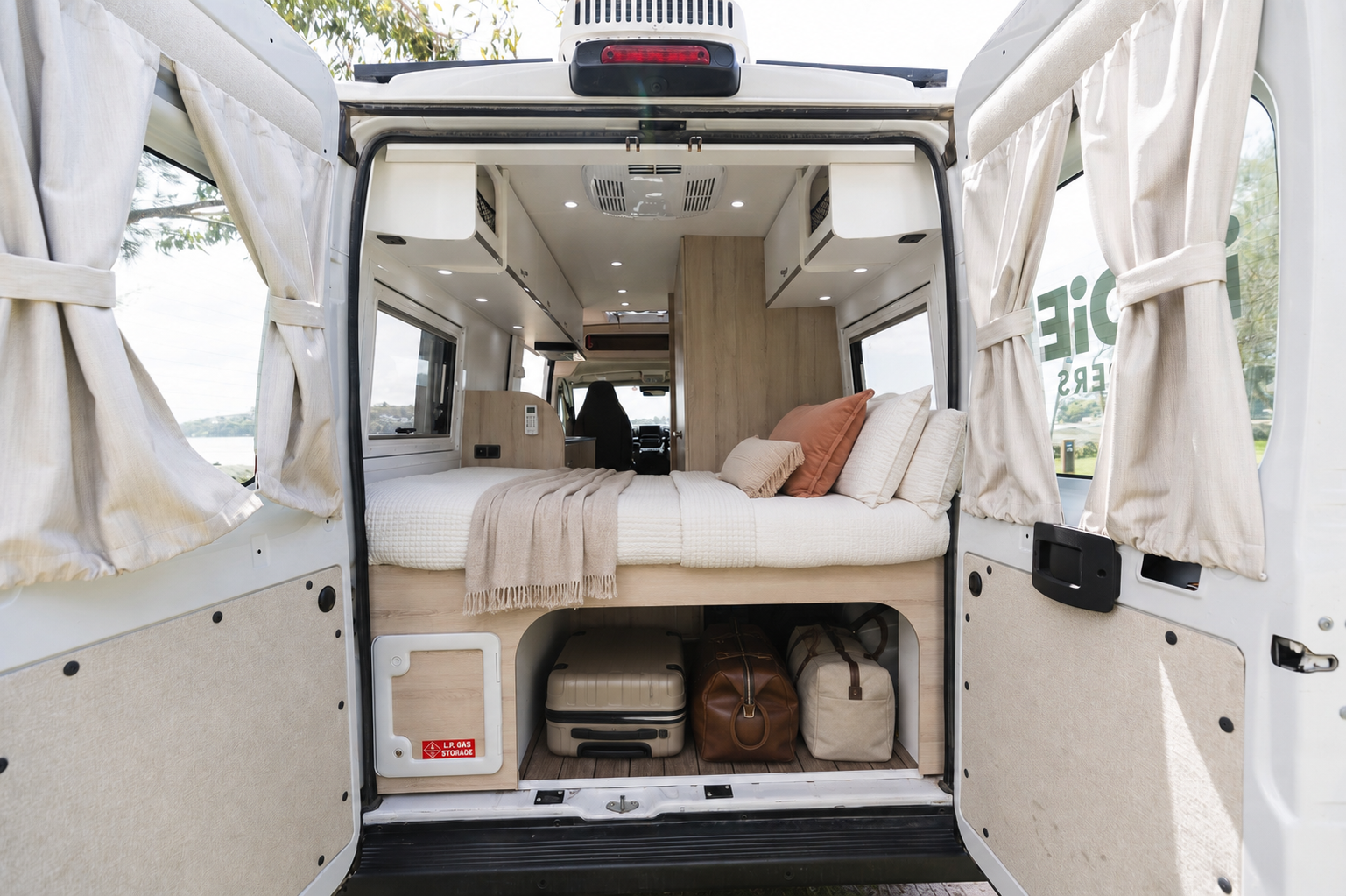 Van interior with rear doors open, showing bed, overhead storage, under-bed luggage space, and light wood furnishings.