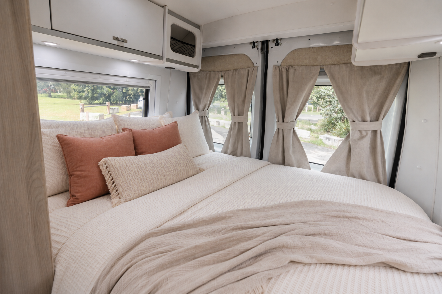 Van interior with double bed, overhead cabinets, pillows, curtains on windows, and light-colored bedding and decor.