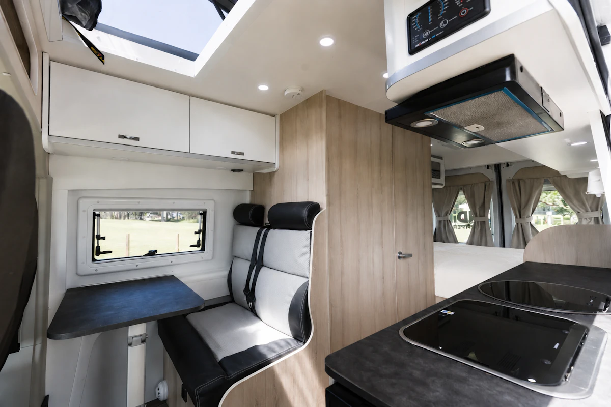 Dining area with two seats, table, overhead cabinets, window, kitchen counter, and bed visible in van interior.
