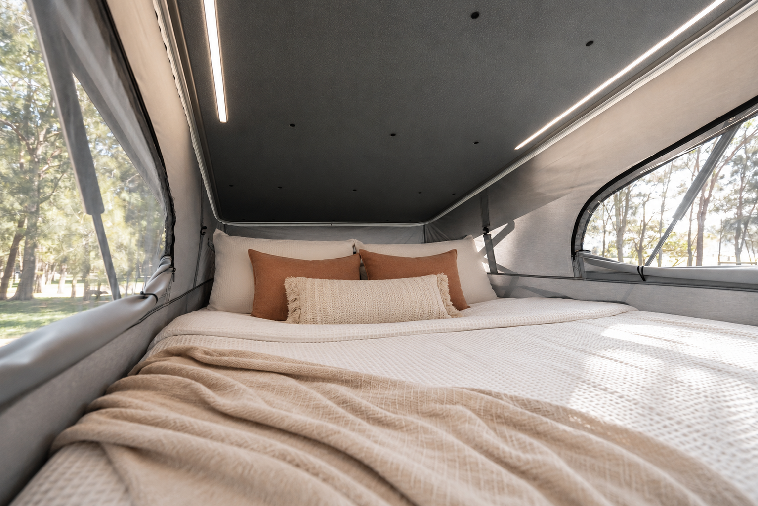 Van interior with made bed, pillows, blanket, windows on both sides, and overhead lighting inside roof tent.