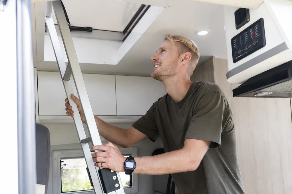 Van interior with ladder, overhead hatch, white cabinets, control panel, and built-in storage visible.