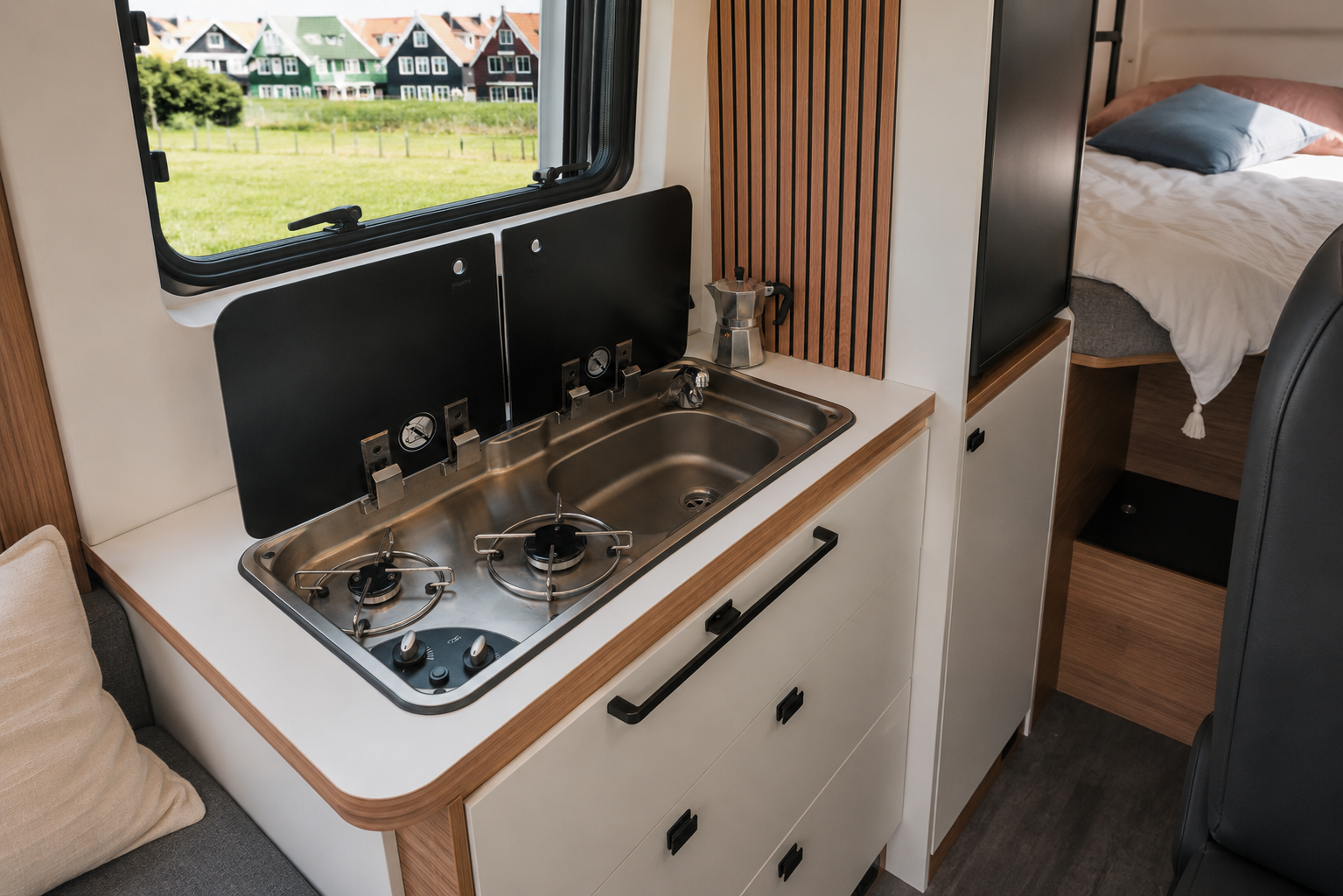 Compact campervan kitchen with two-burner stove, stainless steel sink, counter, cabinets, and window above.