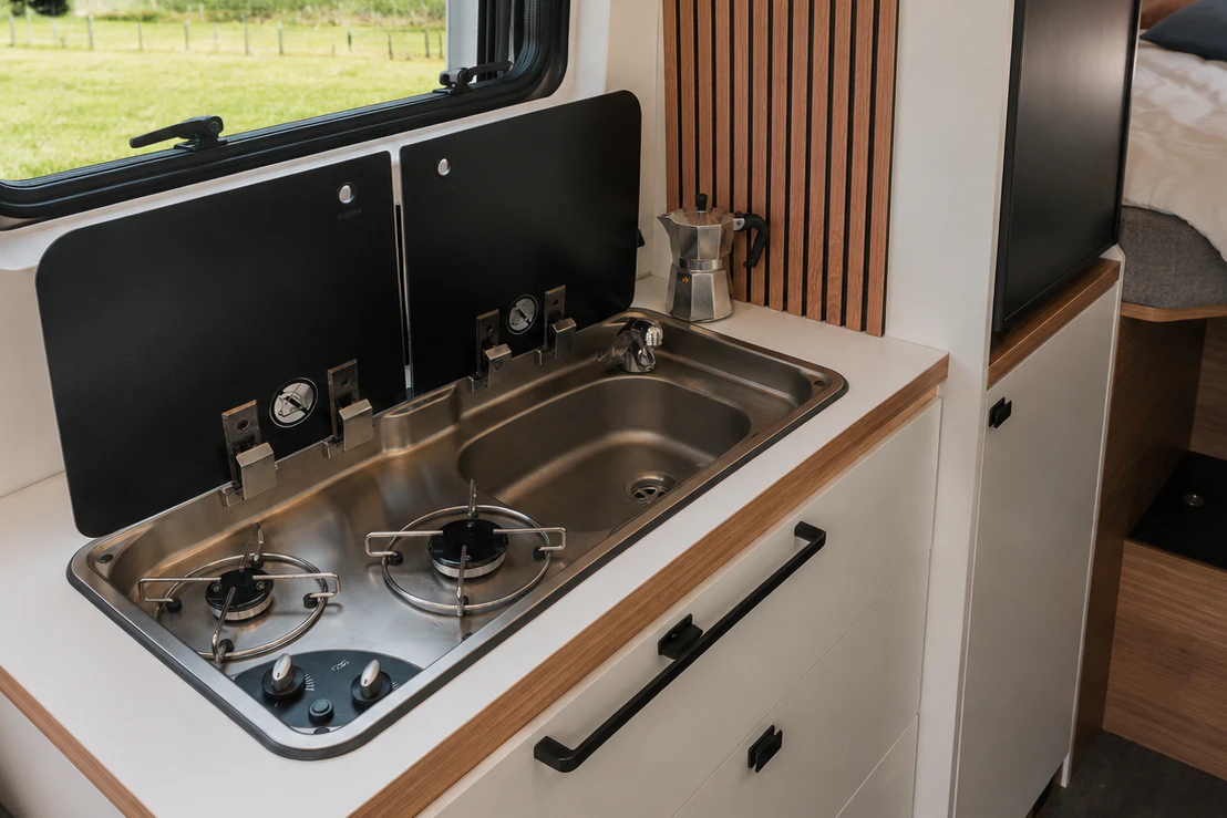 Compact campervan kitchen with two-burner stove, stainless steel sink, counter, cabinets, and window above.