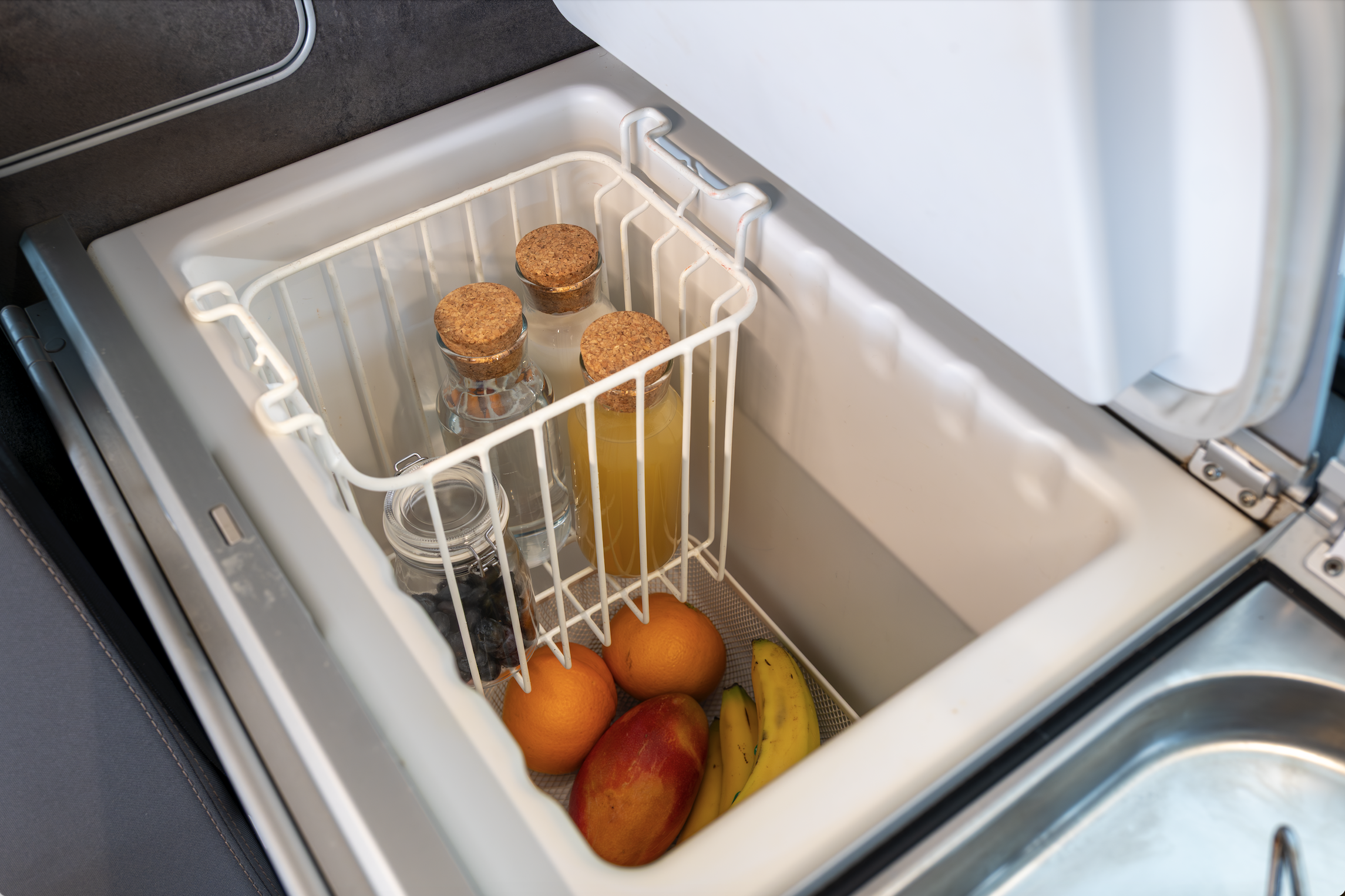 Van interior refrigerator with open lid, containing bottles, bananas, apples, oranges, and a storage basket.