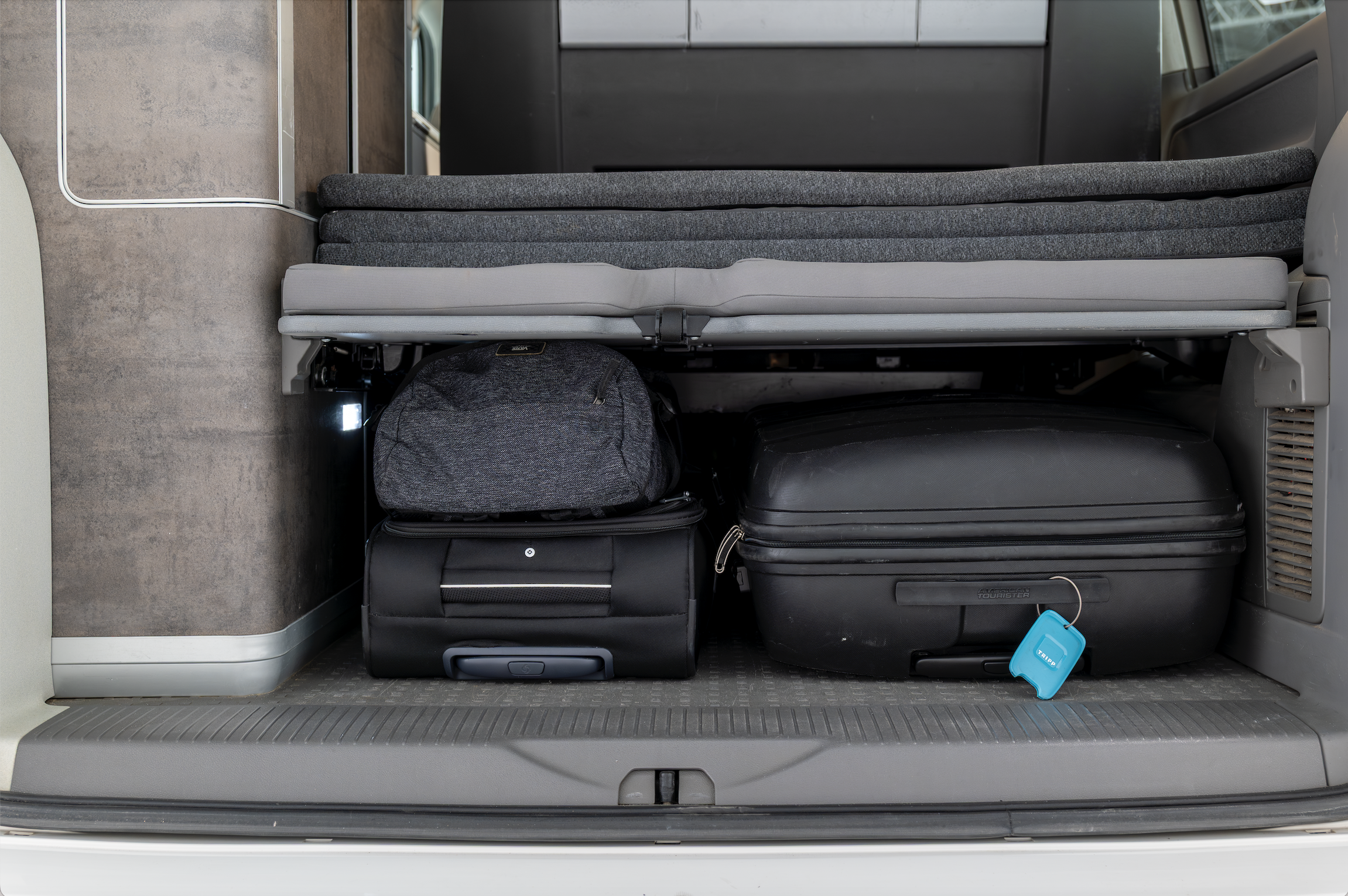 Van interior with folded bed platform above luggage storage area, visible bags and suitcases underneath.