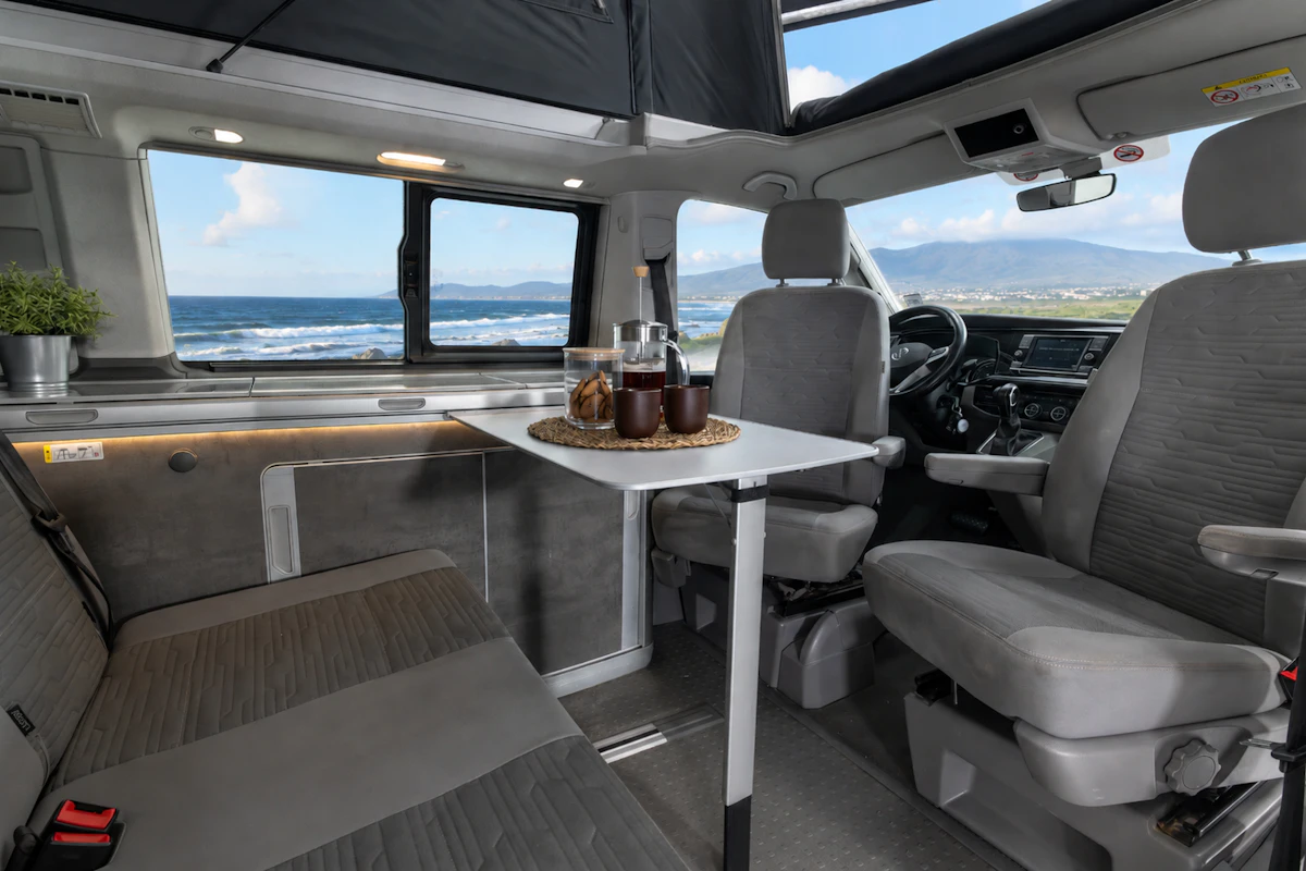 Van interior with front seats, a bench seat, foldable table, small kitchen counter, and window.