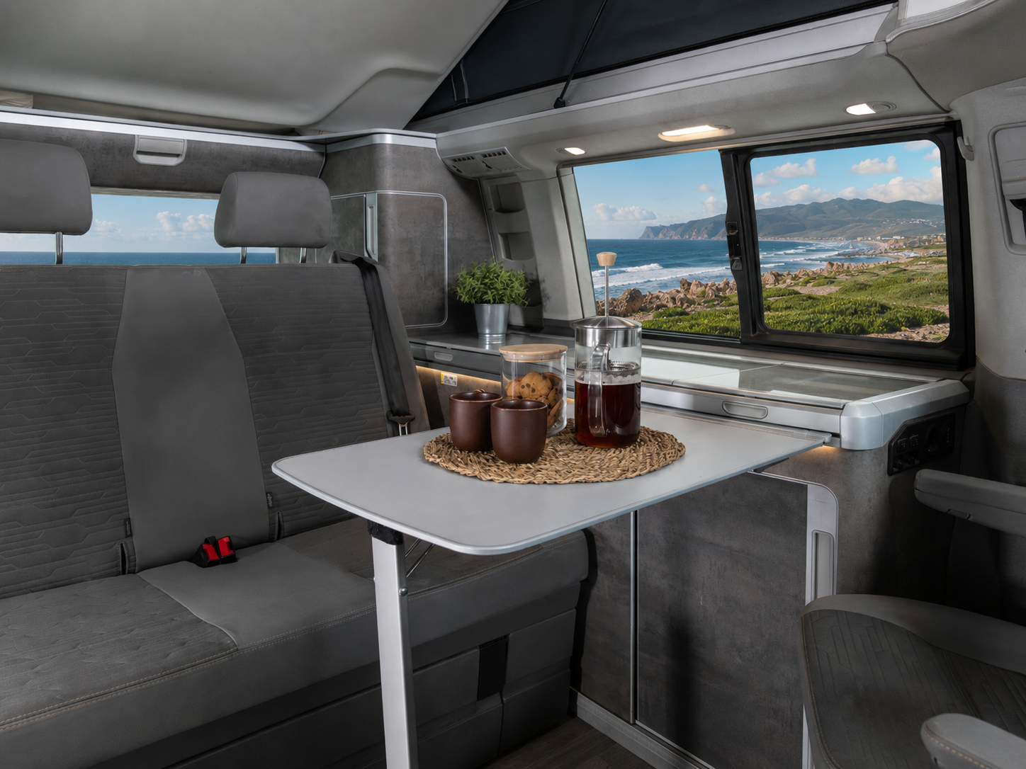 Van interior with grey seats, fold-out table, cups, pitcher, countertop, cabinets, and large side window.