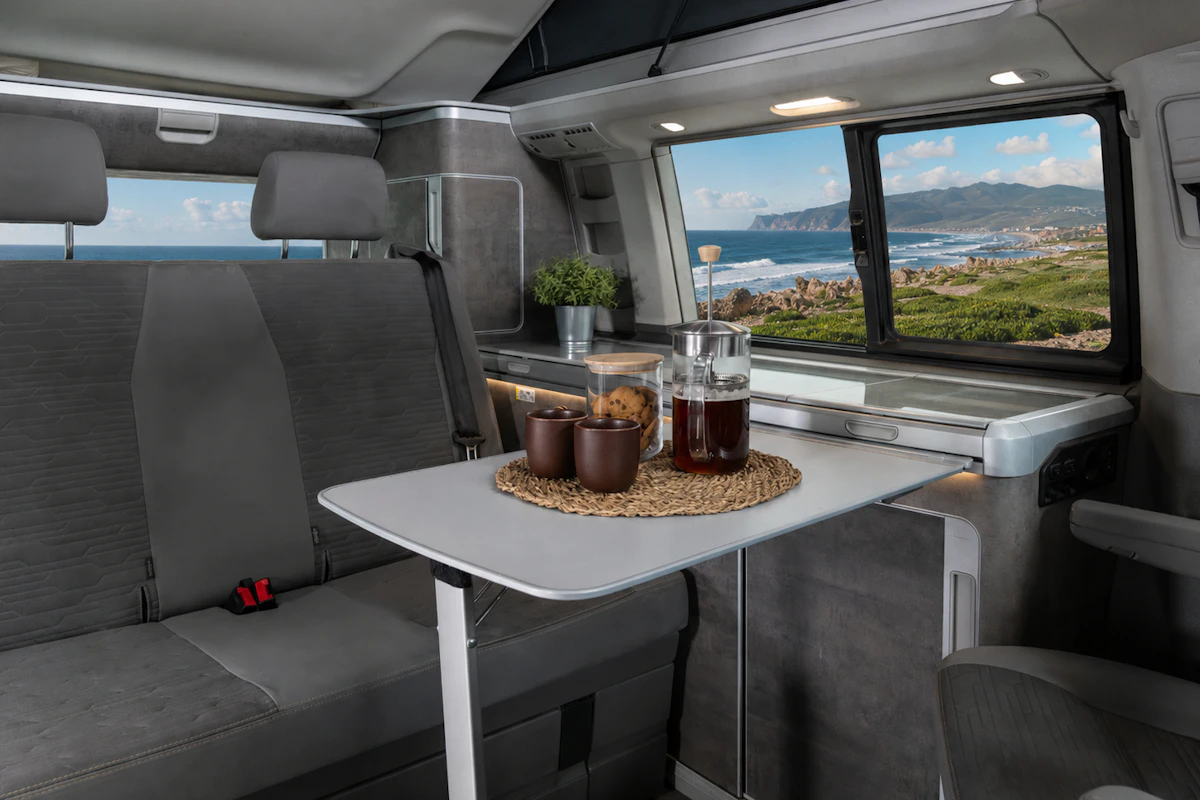 Van interior with grey seats, fold-out table, cups, pitcher, countertop, cabinets, and large side window.