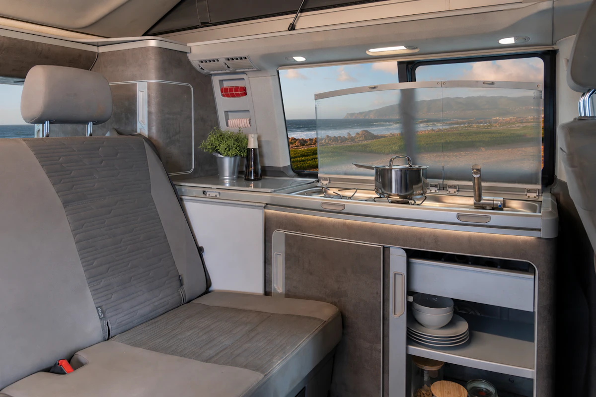 Van interior with two-seat bench, kitchen unit, stove, sink, storage shelves with plates and cups visible.