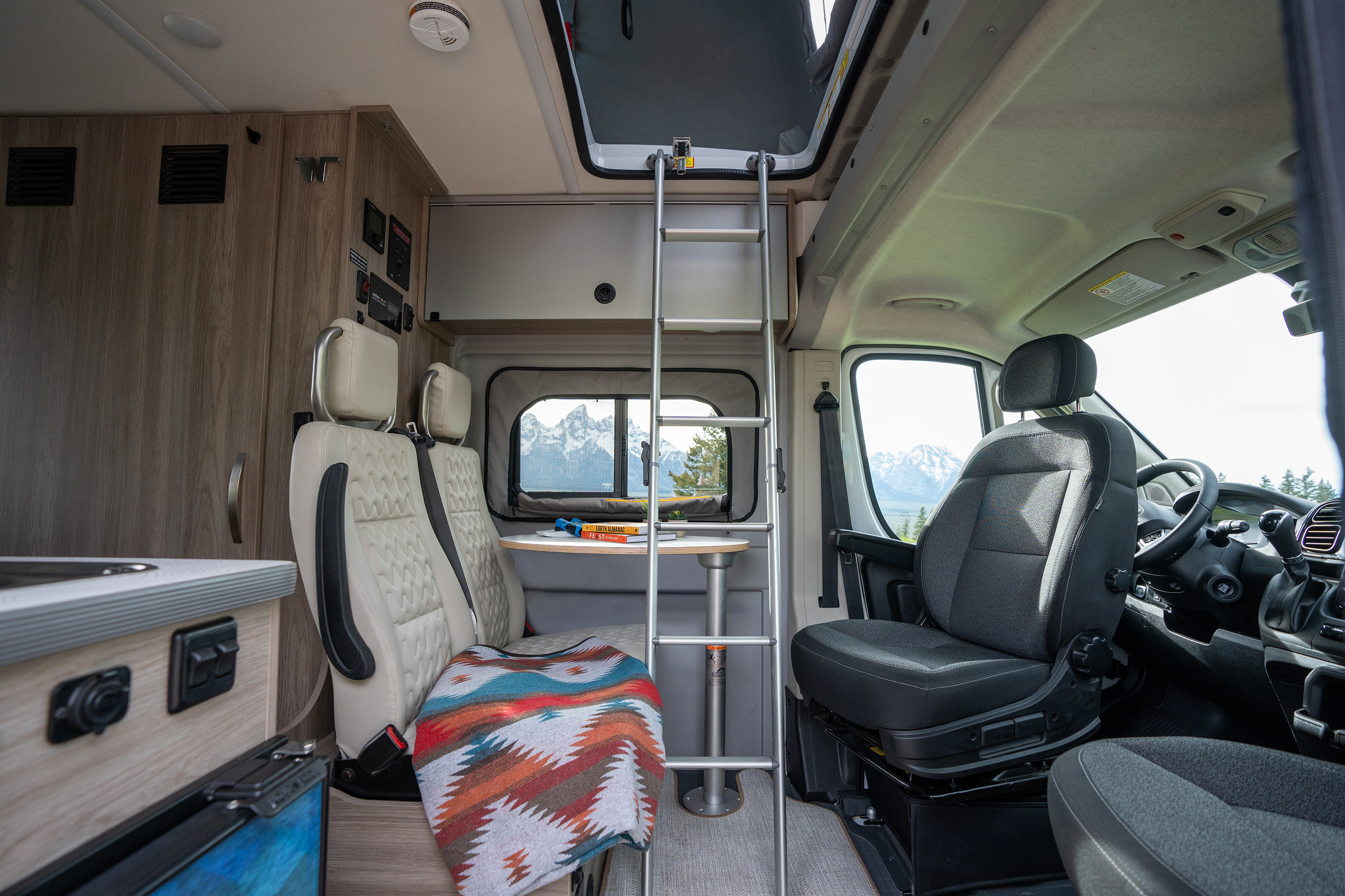 Modern campervan interior with driver and passenger seats, dining area, colorful blanket, loft ladder, and mountain view outside window.