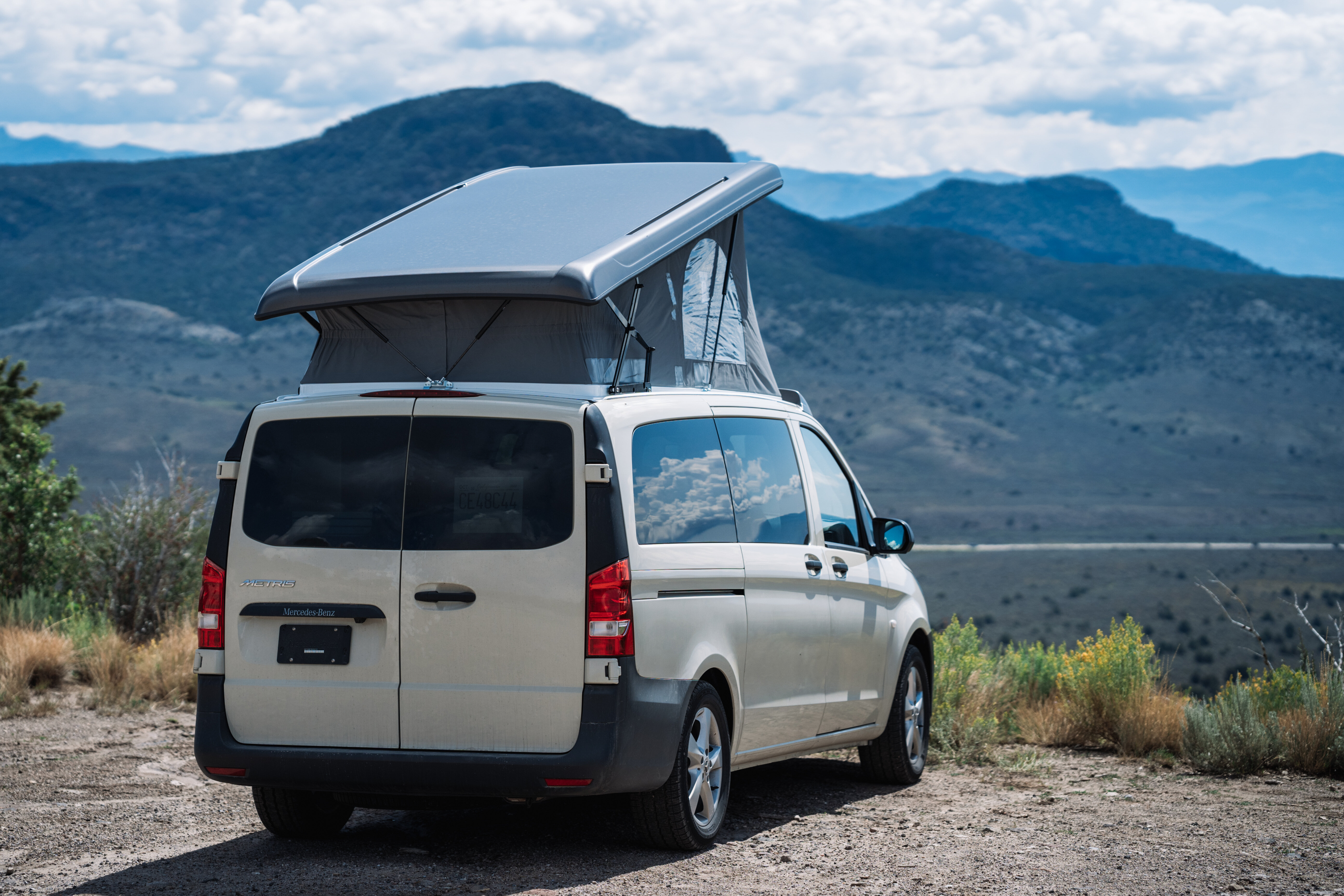 Camper van with pop-up roof parked outdoors, overlooking scenic mountain landscape under a partly cloudy sky.