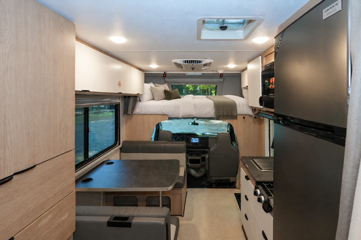 Compact camper van interior with dining table, kitchenette, storage cabinets, upper sleeping area, and front driver’s cab.