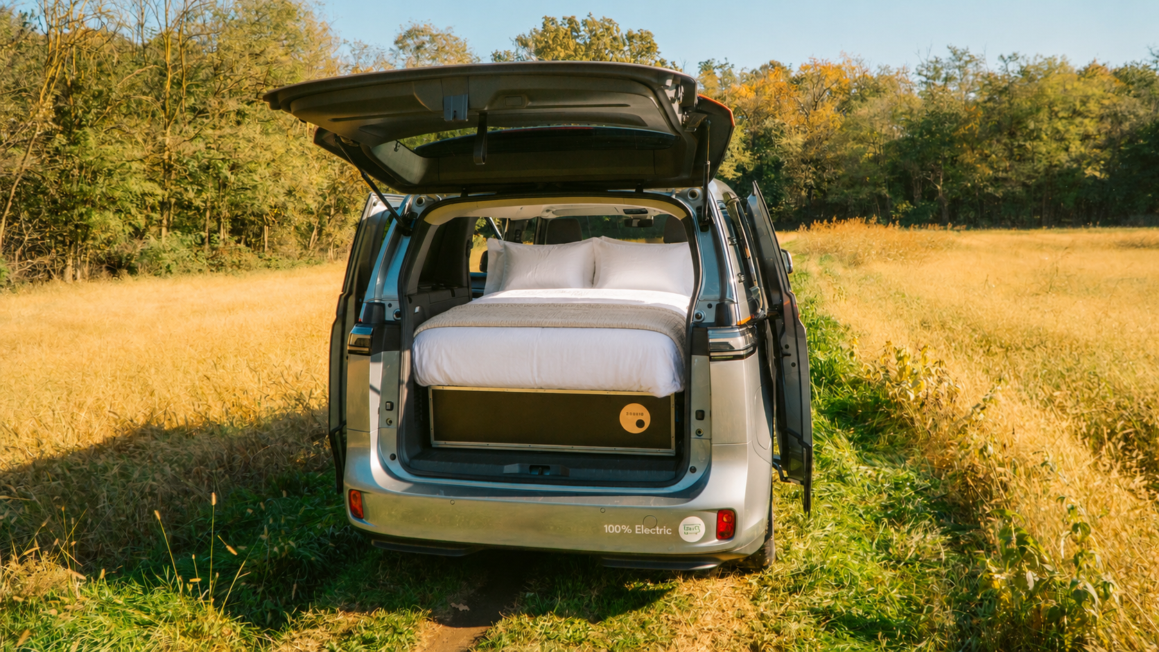 Open trunk of a camper van with a bed setup, parked in a grassy field surrounded by trees.