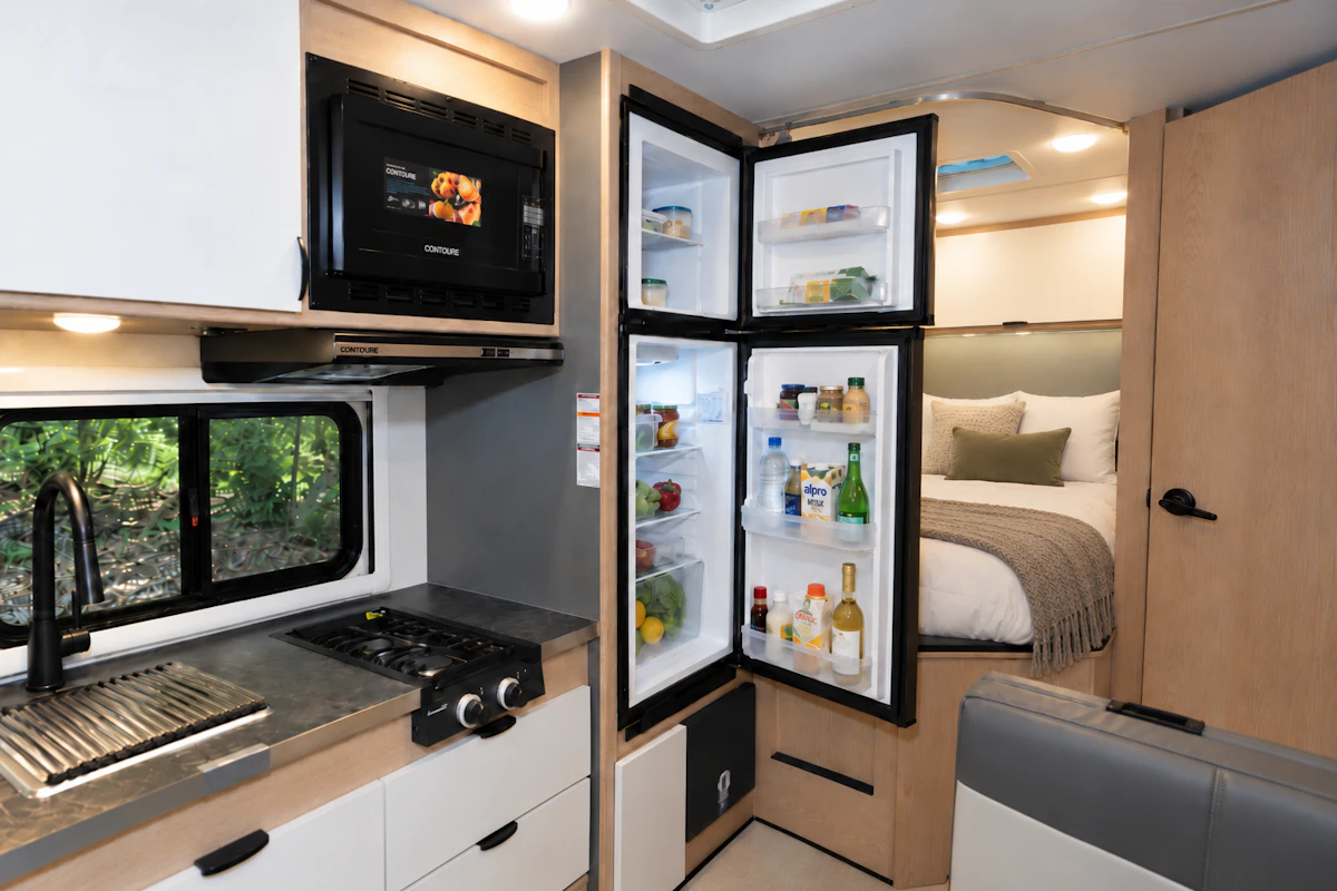 Modern camper van interior with kitchen, open fridge, microwave, sink, stove, and visible rear bed area.