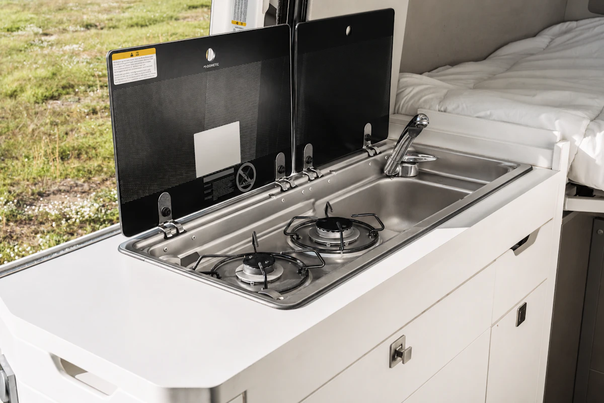 Van interior with countertop, two-burner stove, sink, closed storage, and bed visible in the background.