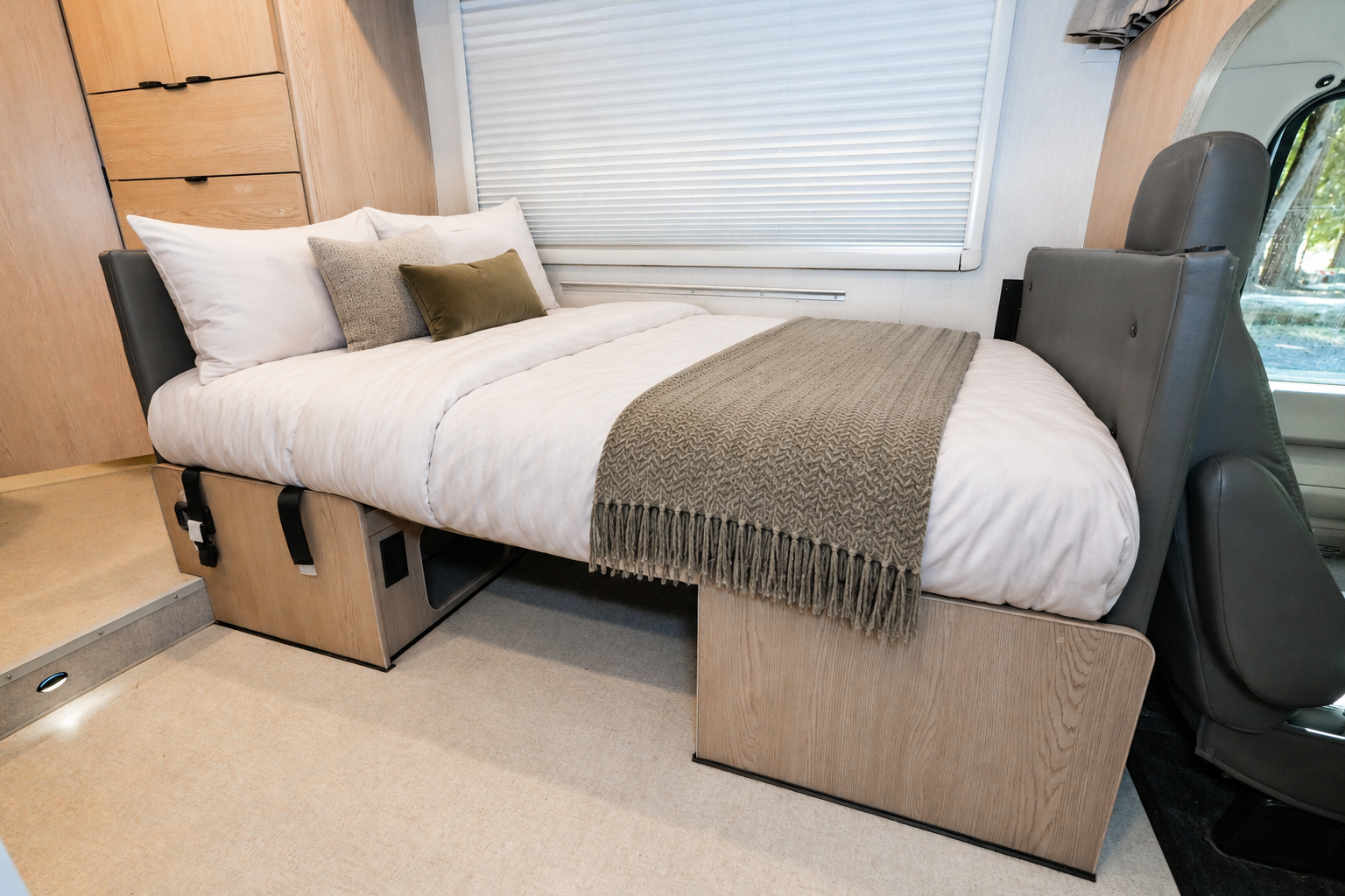 Double bed with pillows, blanket, and storage underneath inside a camper van, adjacent to two swivel seats.