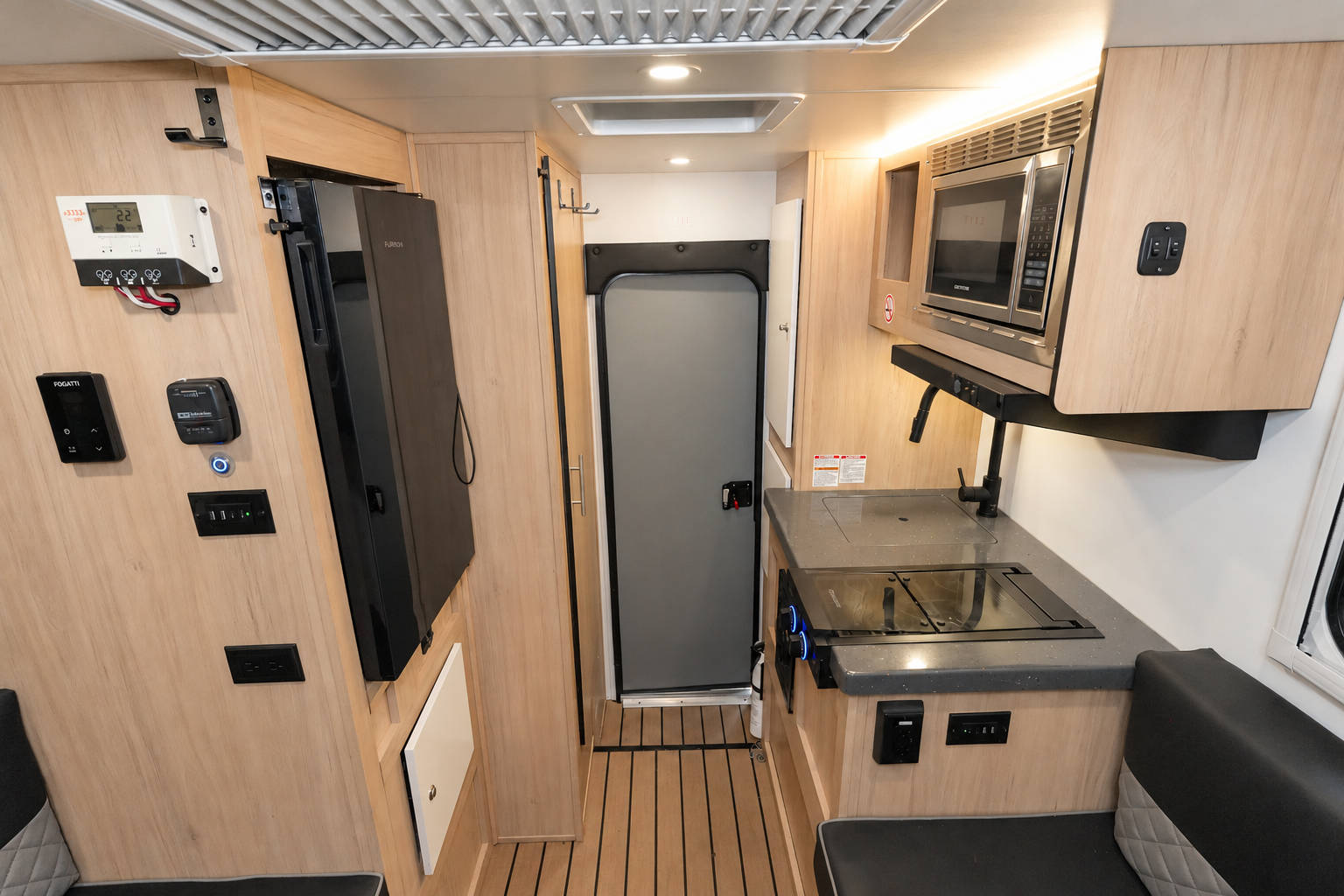 Compact van interior with kitchenette, sink, microwave, cabinets, control panel, and seating area along wood-paneled walls.
