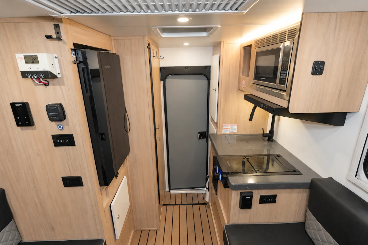 Compact van interior with kitchenette, sink, microwave, cabinets, control panel, and seating area along wood-paneled walls.