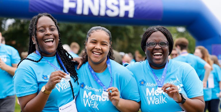 Three ladies wearing #MemoryWalk t-shirts smiling in front of the finish line holding their medals