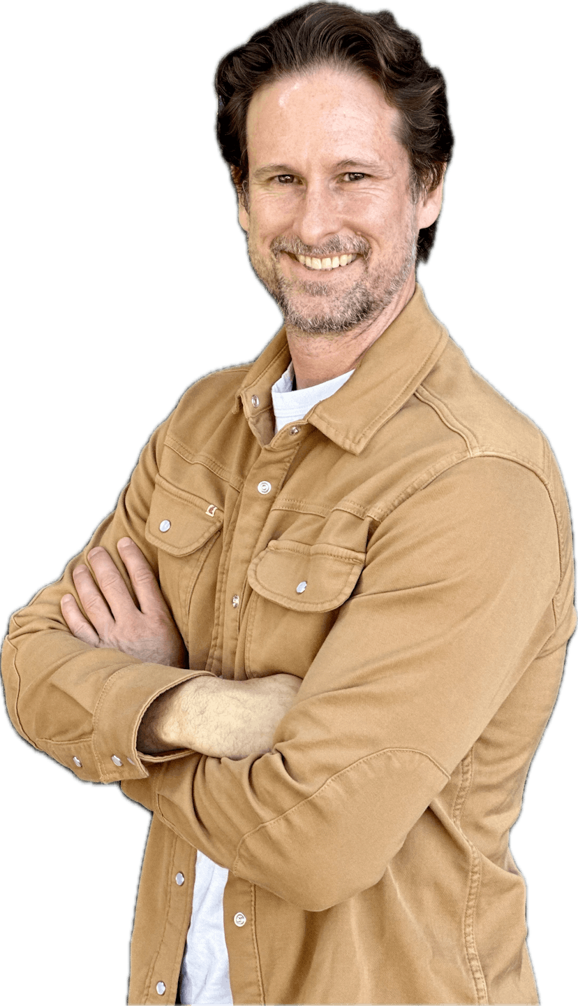 A caucasian man, dressed in a tan overshirt, with dark brown medium length hair, stands smiling with his arms folded.