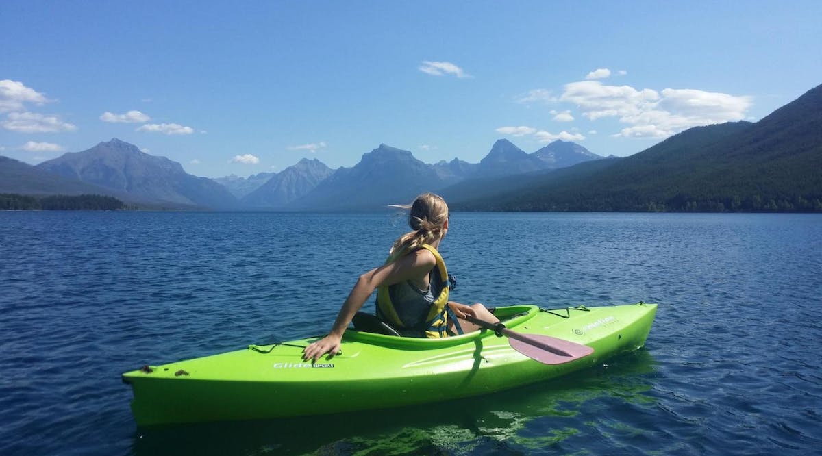 A woman on the water in a green kayak looking towards a vast mountain range.