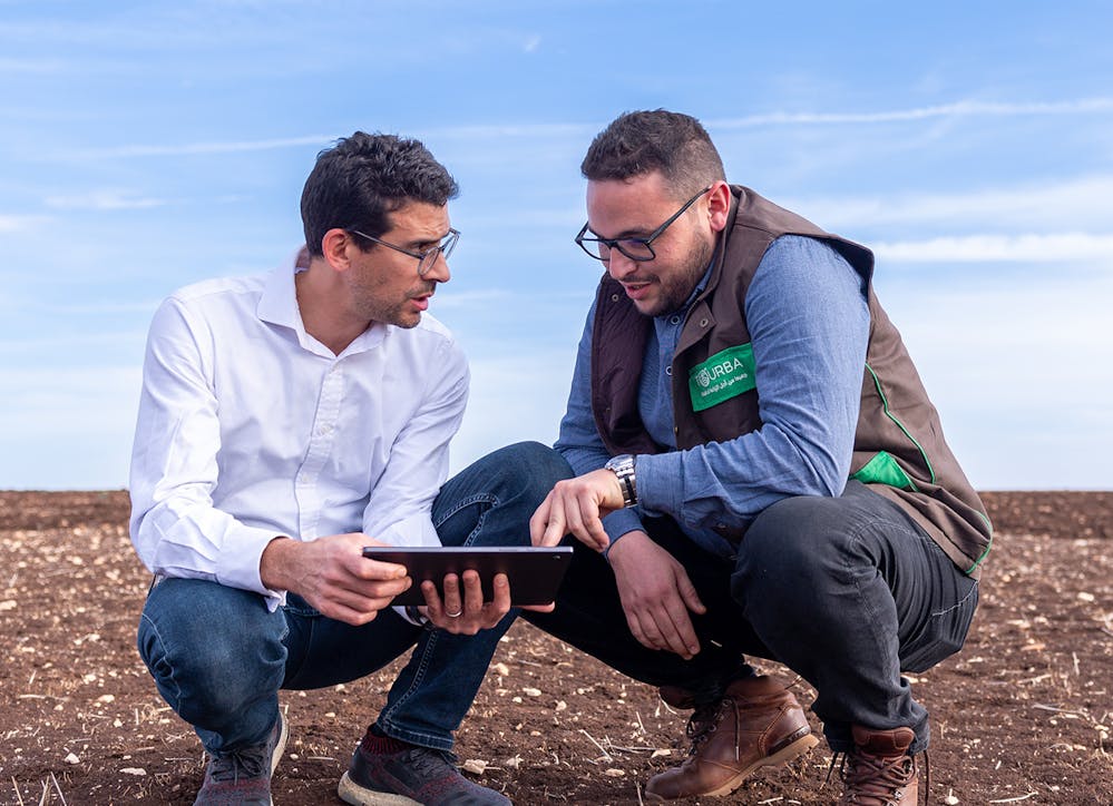 Two professionals kneeling outdoors and reviewing a tablet, representing fieldwork and technical collaboration.