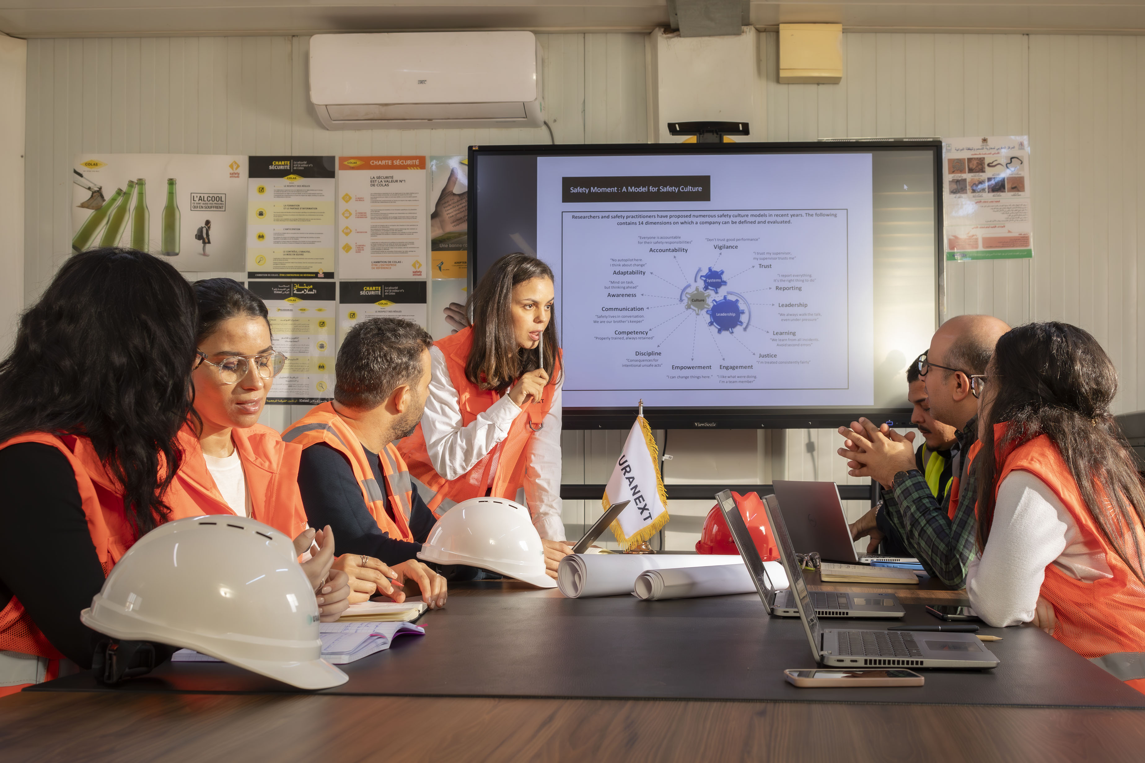 Engineers in safety vests working together in a meeting room, reviewing a technical plan on a screen.