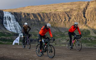 Cyclists are on a dirt road during the Arna Westfjords Way Challenge, with Dynjandi Waterfall in the background. Photo: Photo Gusti Productions