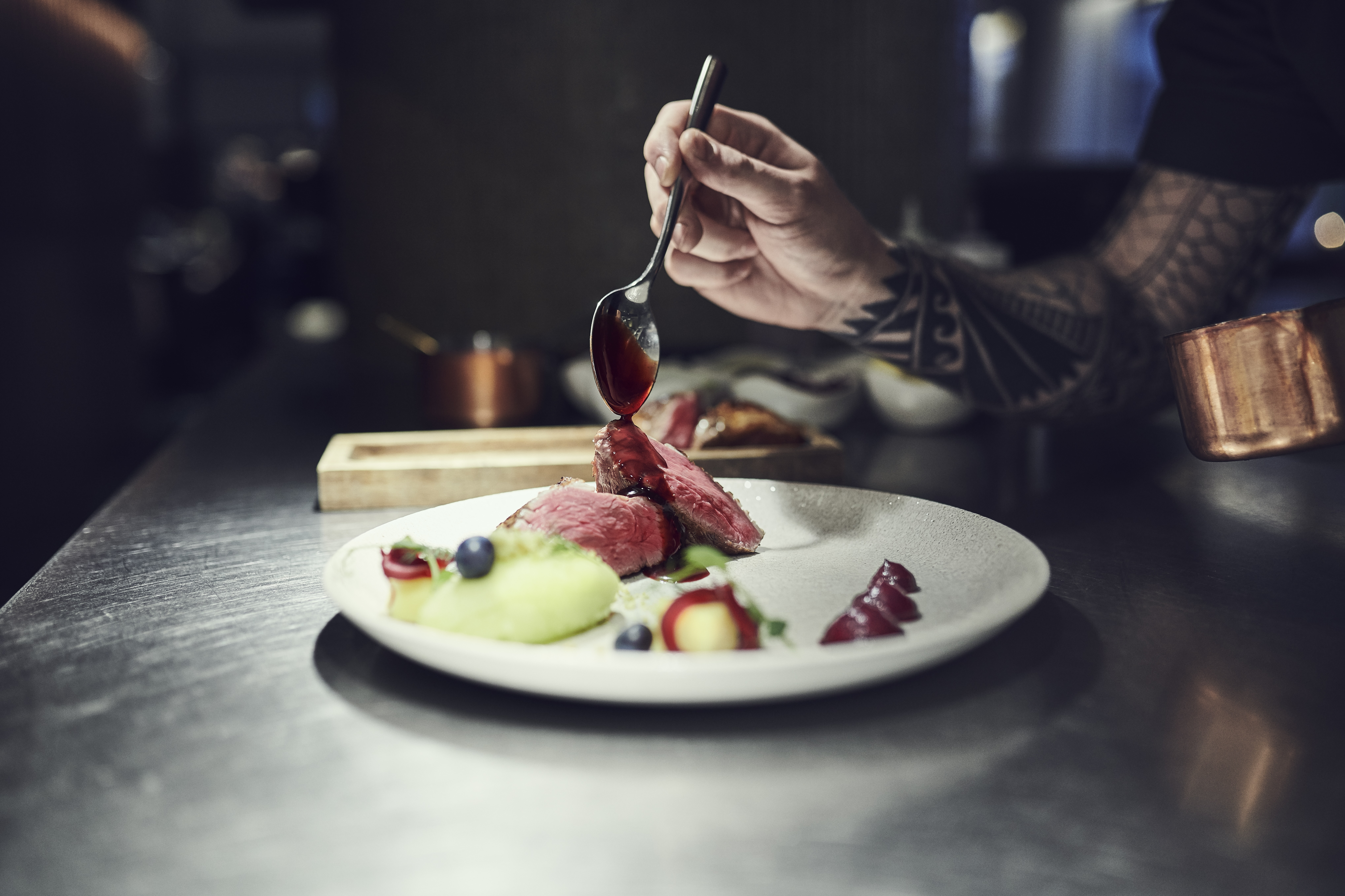 A chef plating a refined Icelandic lamb dish, drizzling sauce over tender slices of meat served with fresh berries and colorful purées on a white plate. Photo: Icelandic Lamb.