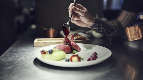 A chef plating a refined Icelandic lamb dish, drizzling sauce over tender slices of meat served with fresh berries and colorful purées on a white plate. Photo: Icelandic Lamb.