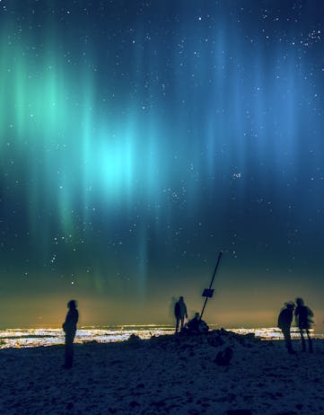 The Northern Lights shimmering above hikers at the top of Úlfarsfell Mountain in the Mosfellsbær city area. 