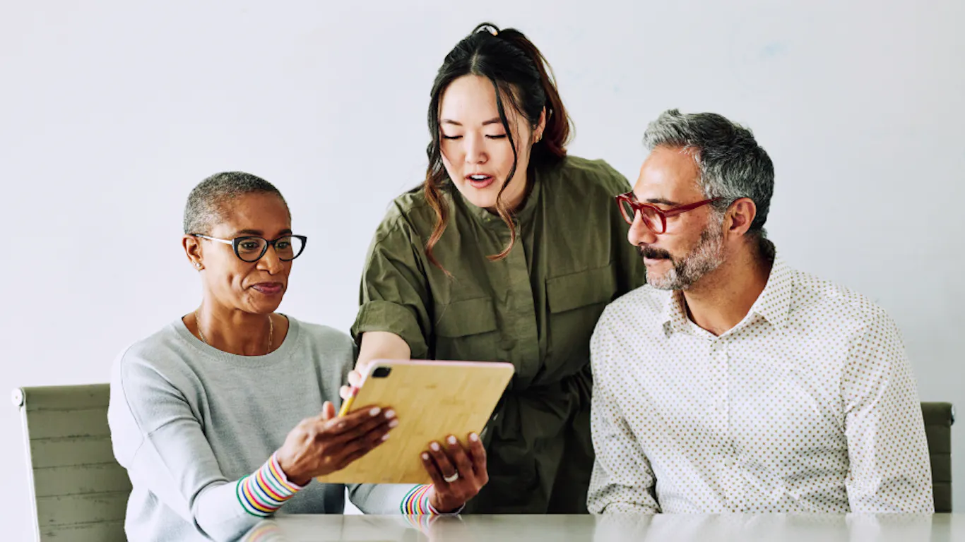 Three people are at a table reviewing a document. Two people are sitting; one person is standing behind them, pointing to a document being held by one of the people sitting at the table. 