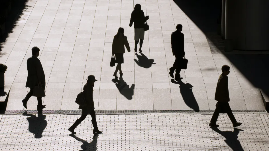 People are walking in the sun on a tiled surface. The sunlight is bright, and the people are cast in a dark shadow.