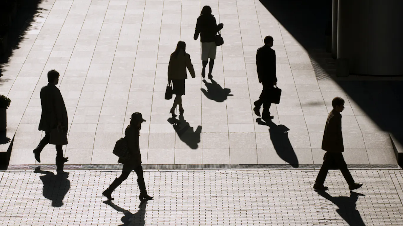 People are walking in the sun on a tiled surface. The sunlight is bright, and the people are cast in a dark shadow. 