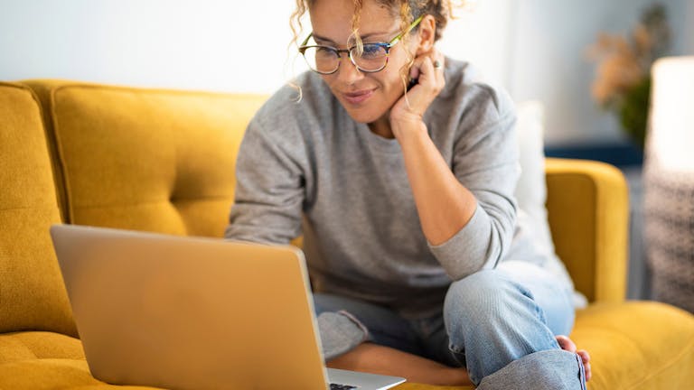 A woman sitting on a couch, smiling and looking at her laptop