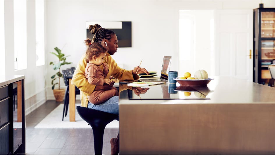 woman holding her child and working on her laptop at home