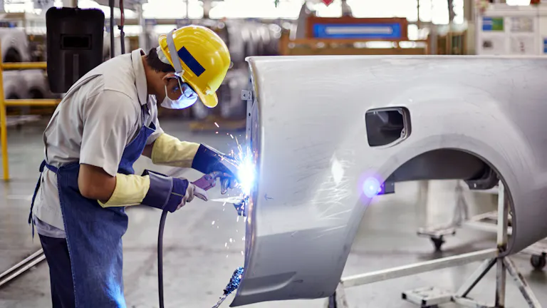 A person welding on a large part that will be added to a car build. The part is white and includes a wheel well.