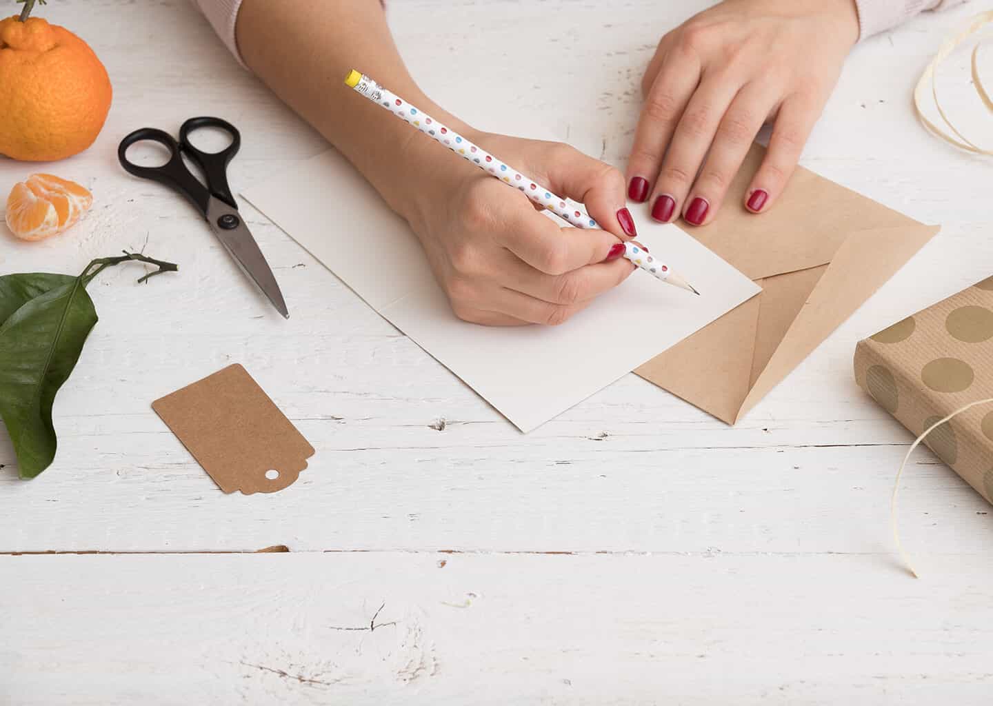 Yakın plan çekimde, ahşap masa üzerinde kadın düğün davetiyesine yazı yazıyor. Masa üzerinde davetiye zarfı, makas, hediye paketi ve mandalina bulunuyor.