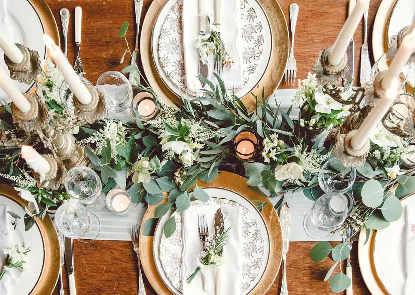 Ahşap yemek masası üzerinde şamdanlar, tabaklar, çatallar, bıçaklar ve yapraklar bulunuyor.